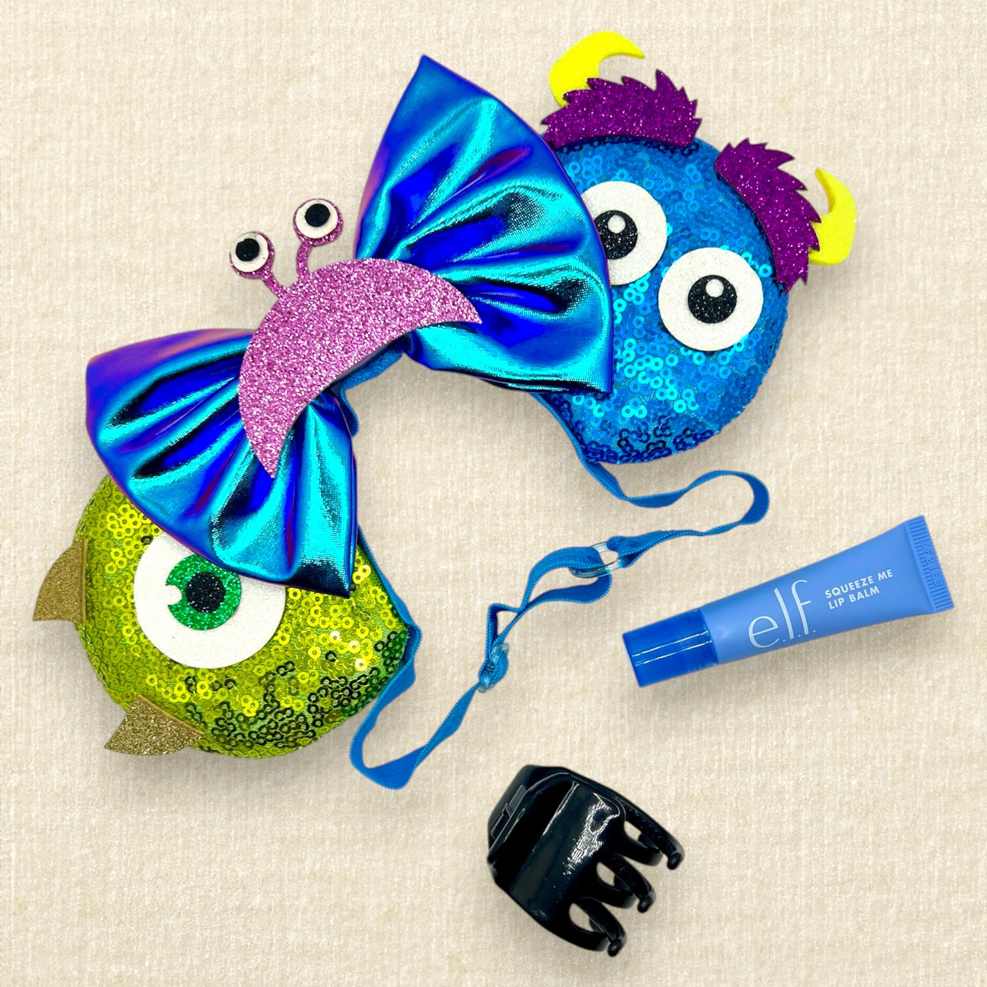 Green and blue sequin mouse ears with glitter monster details and an iridescent blue bow on an elastic headband; black claw clip; tube of lip balm