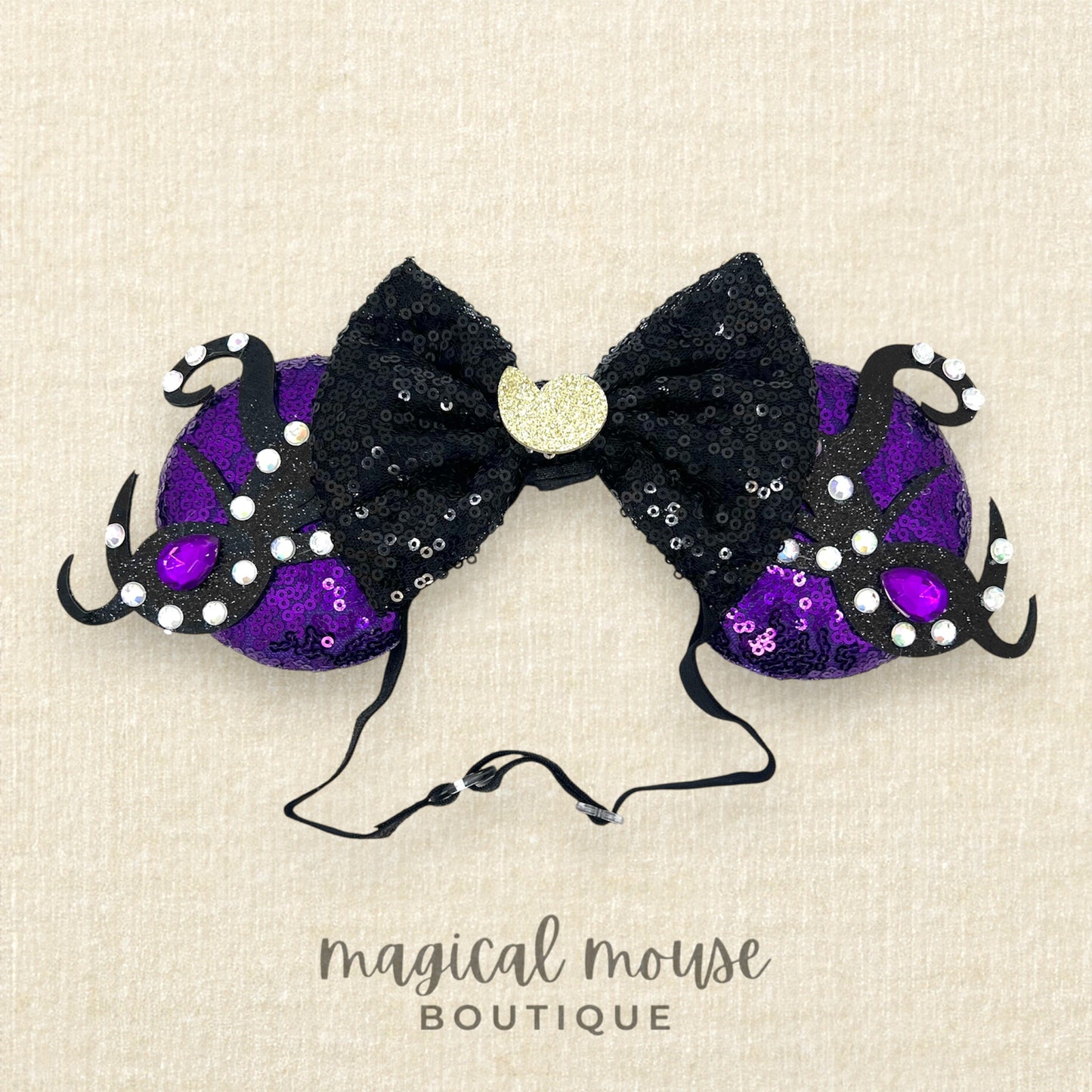 Black and purple mouse ears headband with a black bow, glitter silver seashell centerpiece, black glitter tentacles, and purple and crystal rhinestones on a beige background, branded 'magical mouse boutique'.