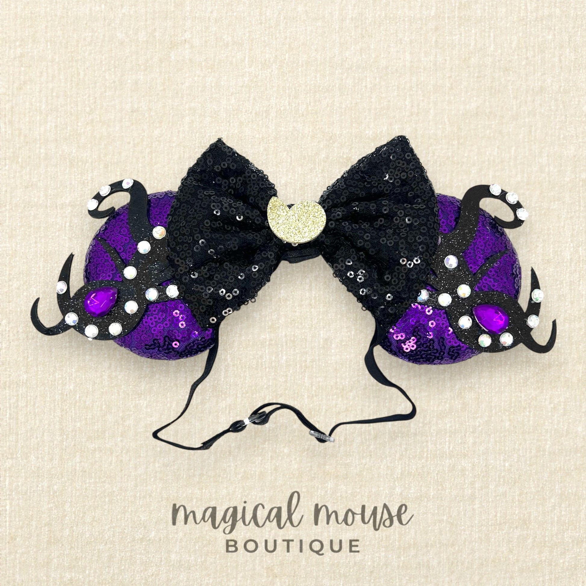 Black and purple mouse ears headband with a black bow, glitter silver seashell centerpiece, black glitter tentacles, and purple and crystal rhinestones on a beige background, branded 'magical mouse boutique'.