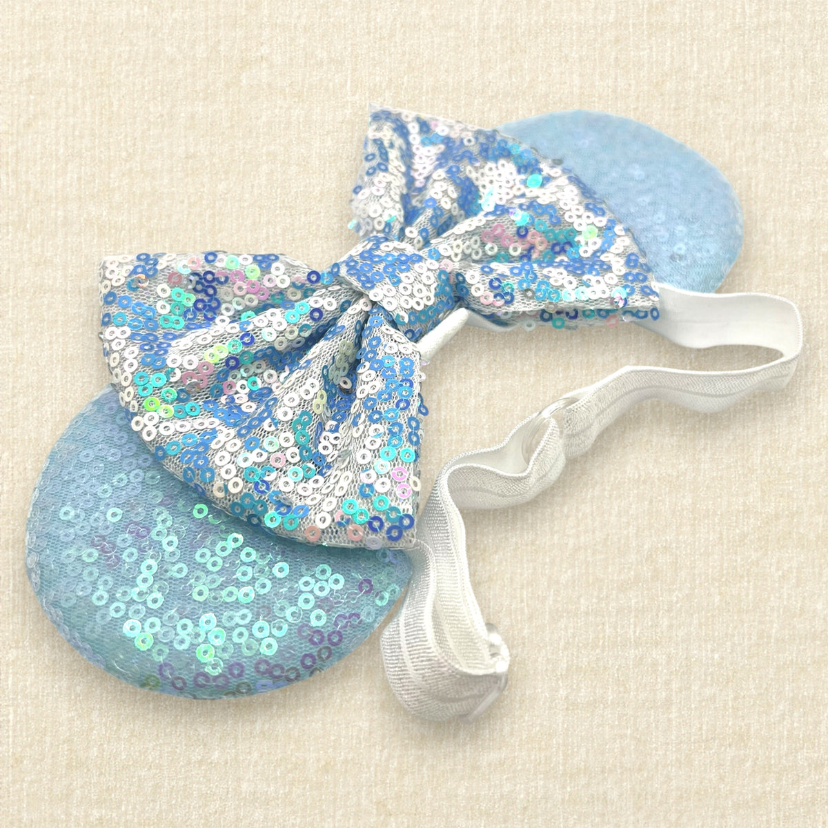 A winter-themed mouse ears headband with iridescent light blue sequin mouse ears and a blue and white sequin bow on an elastic headband
