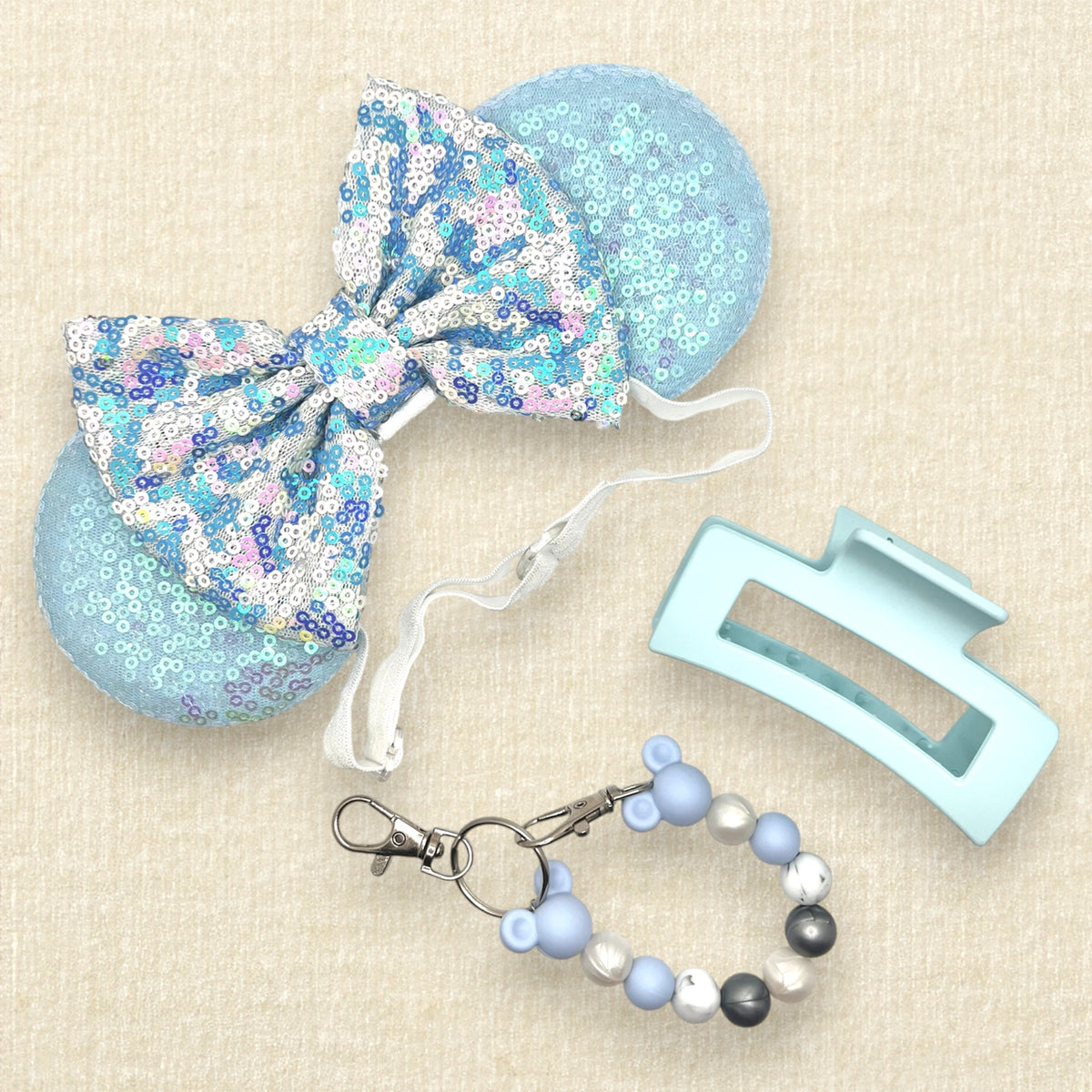 A winter-themed mouse ears headband with iridescent light blue sequin mouse ears and a blue and white sequin bow on an elastic headband; a blue, pearl white, and silver beaded mouse ears holder keychain clip; and a light blue claw clip
