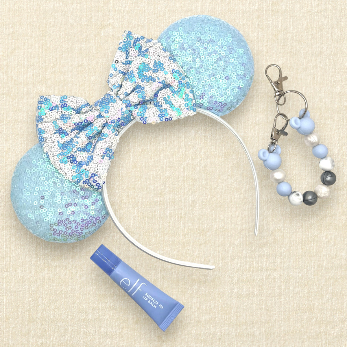 A winter-themed mouse ears headband with iridescent light blue sequin mouse ears and a blue and white sequin bow; blue tube of lip gloss; and a blue, pearl white, and silver beaded mouse ears holder keychain clip