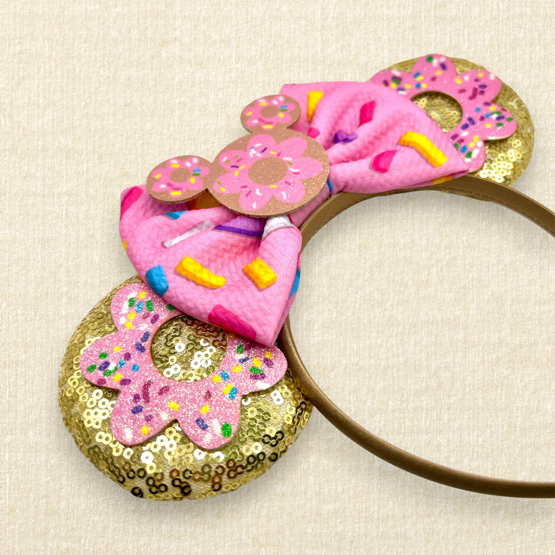 Gold mouse ears with pink glitter sprinkle donuts, a pink fabric bow with rainbow sprinkles, and a mouse-shaped donut centerpiece