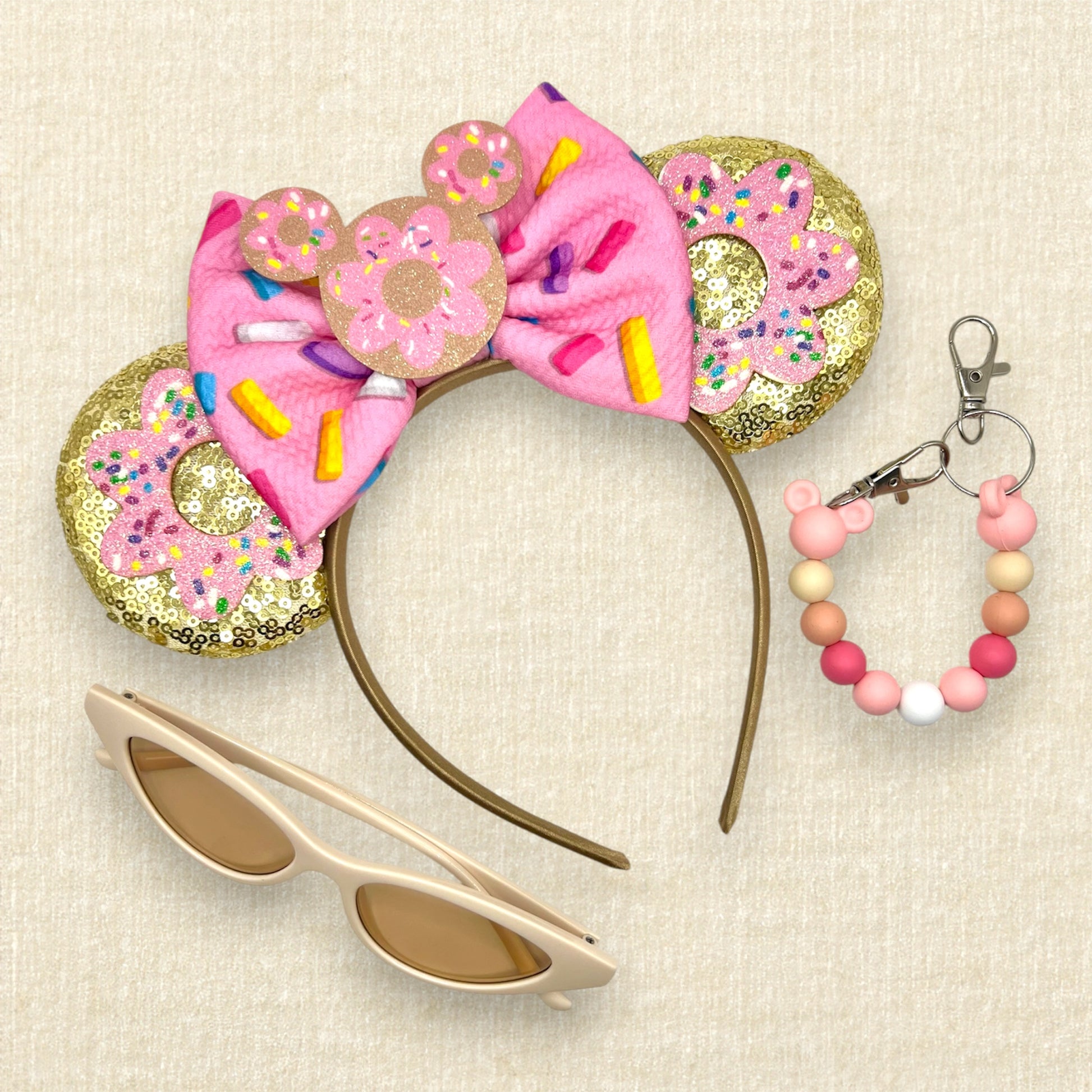 Gold mouse ears with pink glitter sprinkle donuts, a pink fabric bow with rainbow sprinkles, and a mouse-shaped donut centerpiece; cream sunglasses; pink beaded mouse ears holder