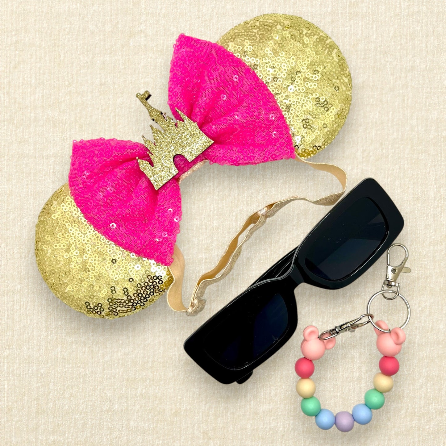 Gold sequin mouse ears with hot pink sequin bow and gold glitter storybook castle on an elastic headband; black sunglasses; colorful rainbow beaded mouse ears holder keychain
