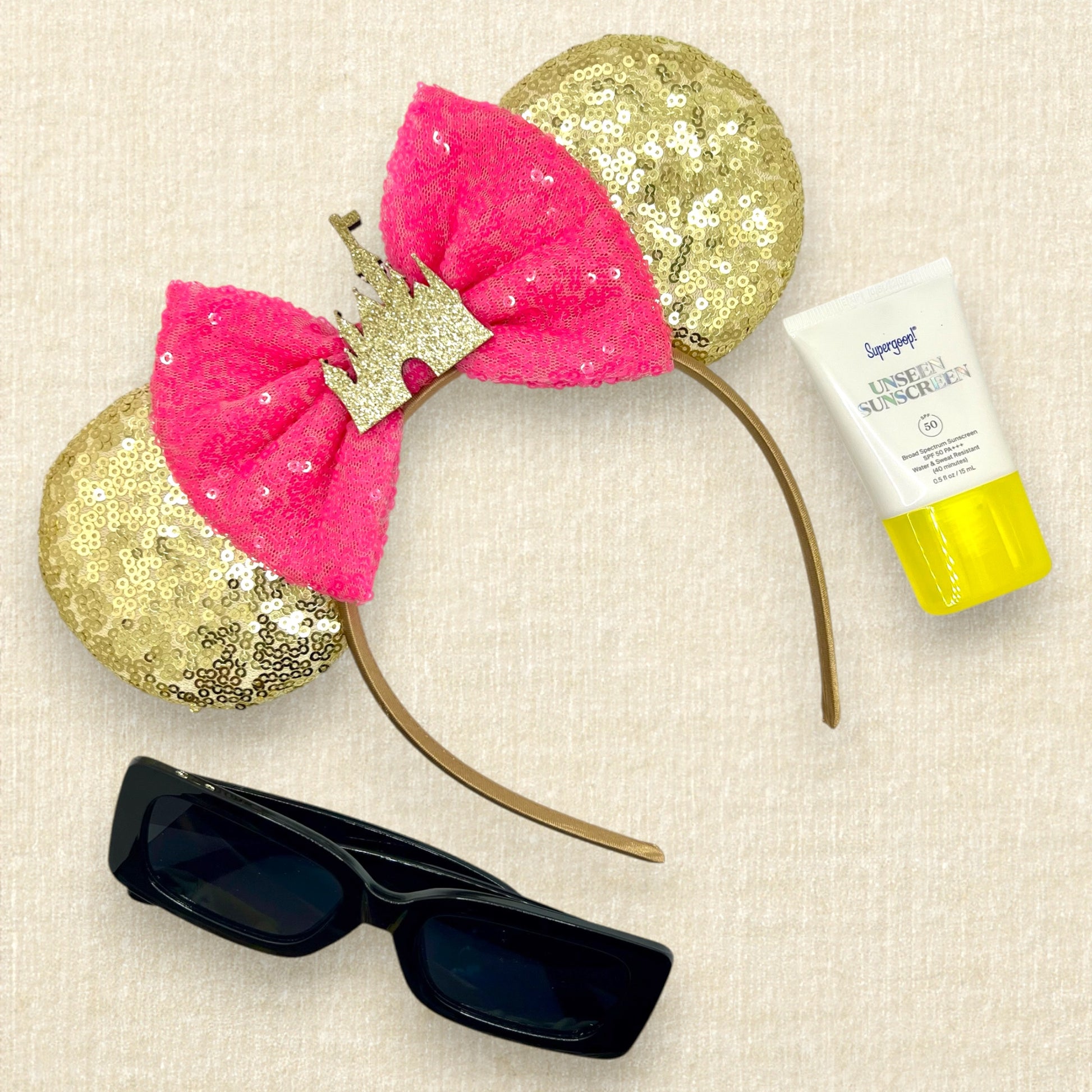 Gold sequin mouse ears with hot pink sequin bow and gold glitter castle; black sunglasses; small tube of sunscreen