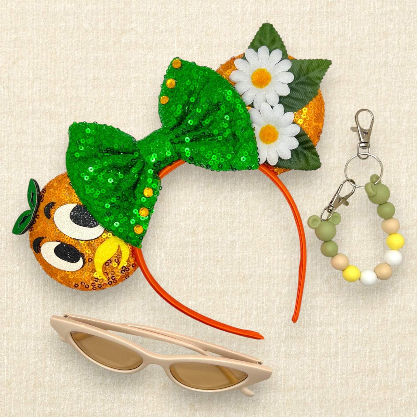 Orange sequin mouse ears with glitter bird details, white fabric daisies, and green sequin bow; cream cat-eye sunglasses; green and yellow beaded mouse ears holder keychain