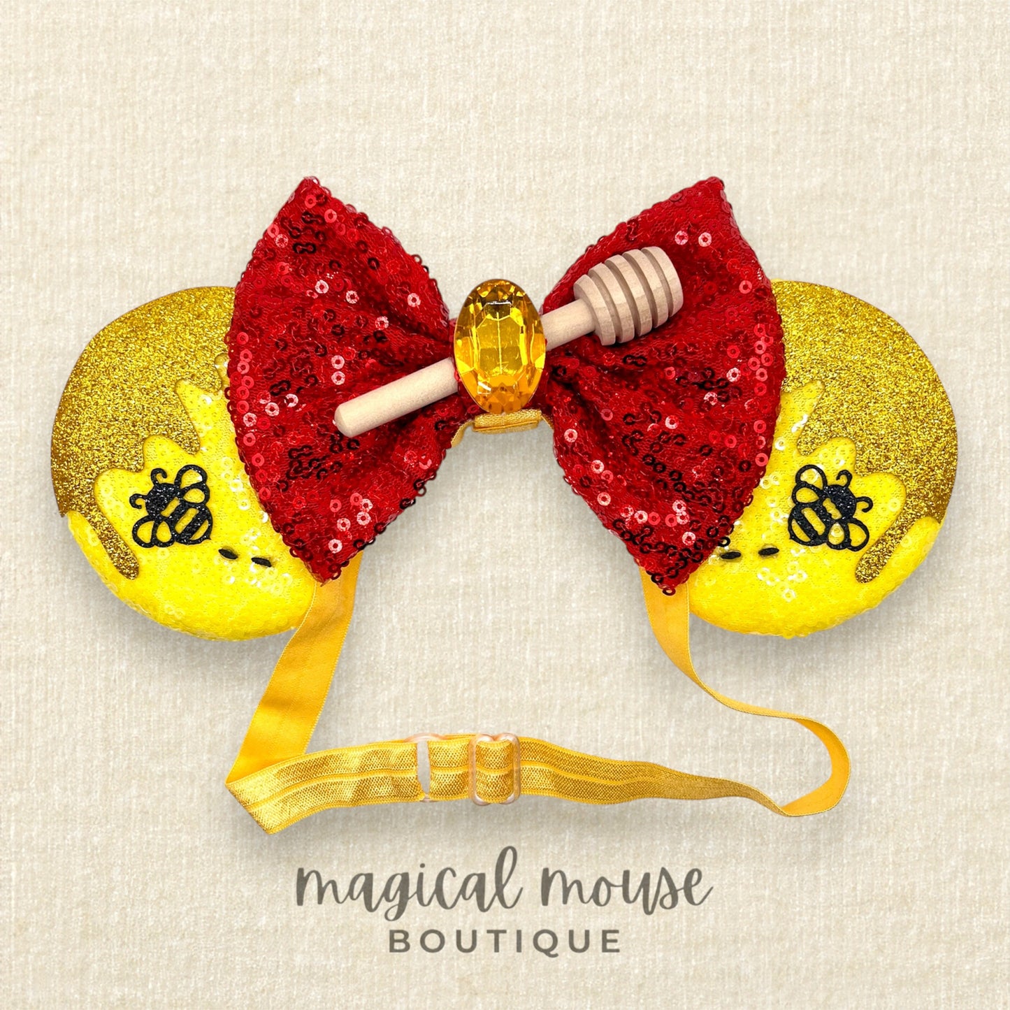 Decorative mouse ears headband with red sequin bow, yellow ears, and honeycomb design on a beige background. Brand 'magical mouse boutique' visible.