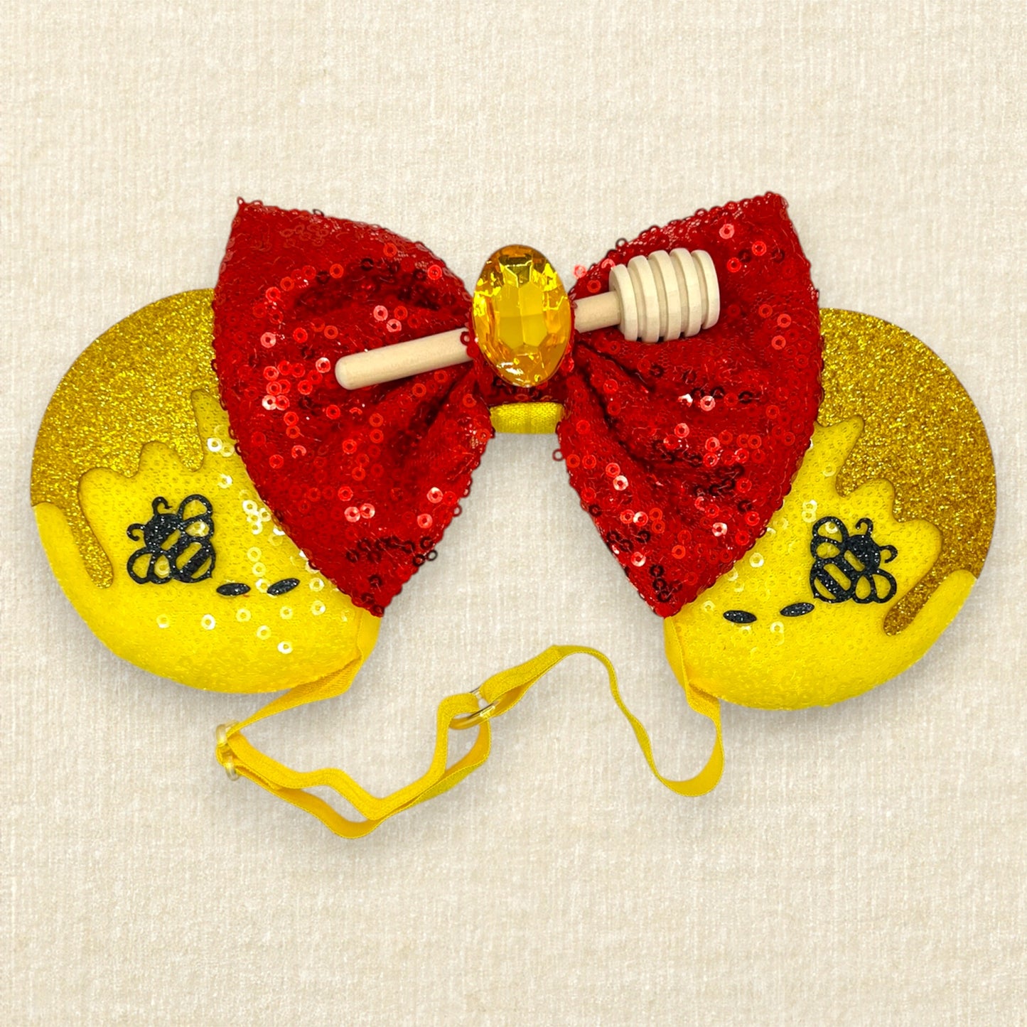 Yellow sequin mouse ears with glitter honey and bee accents, red sequin bow, wooden honey dipper, and yellow rhinestone on an elastic headband