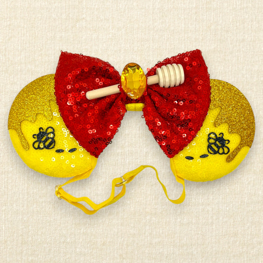 Yellow sequin mouse ears with glitter honey and bee accents, red sequin bow, wooden honey dipper, and yellow rhinestone on an elastic headband