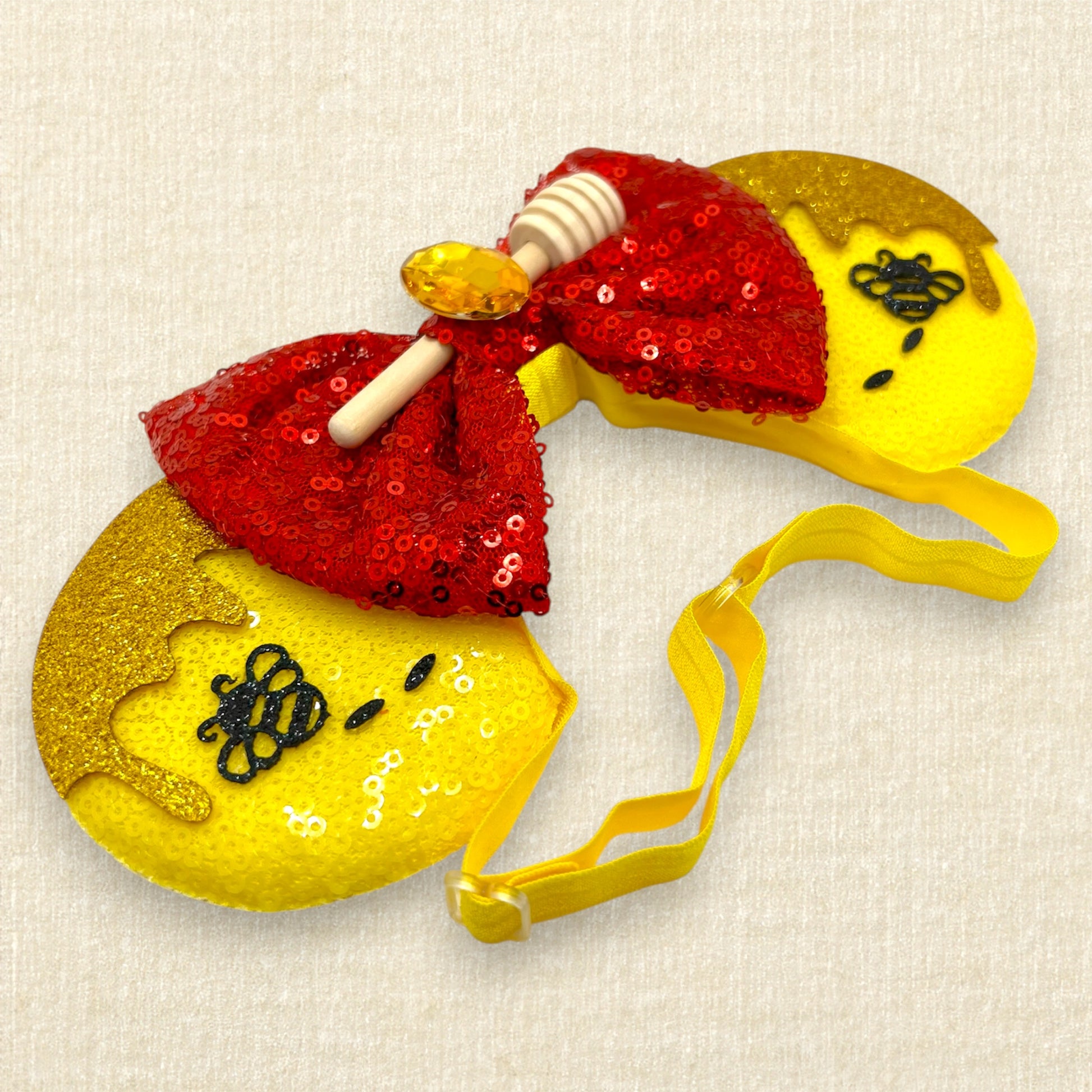 Yellow sequin mouse ears with glitter honey and bee accents, red sequin bow, wooden honey dipper, and yellow rhinestone on an elastic headband