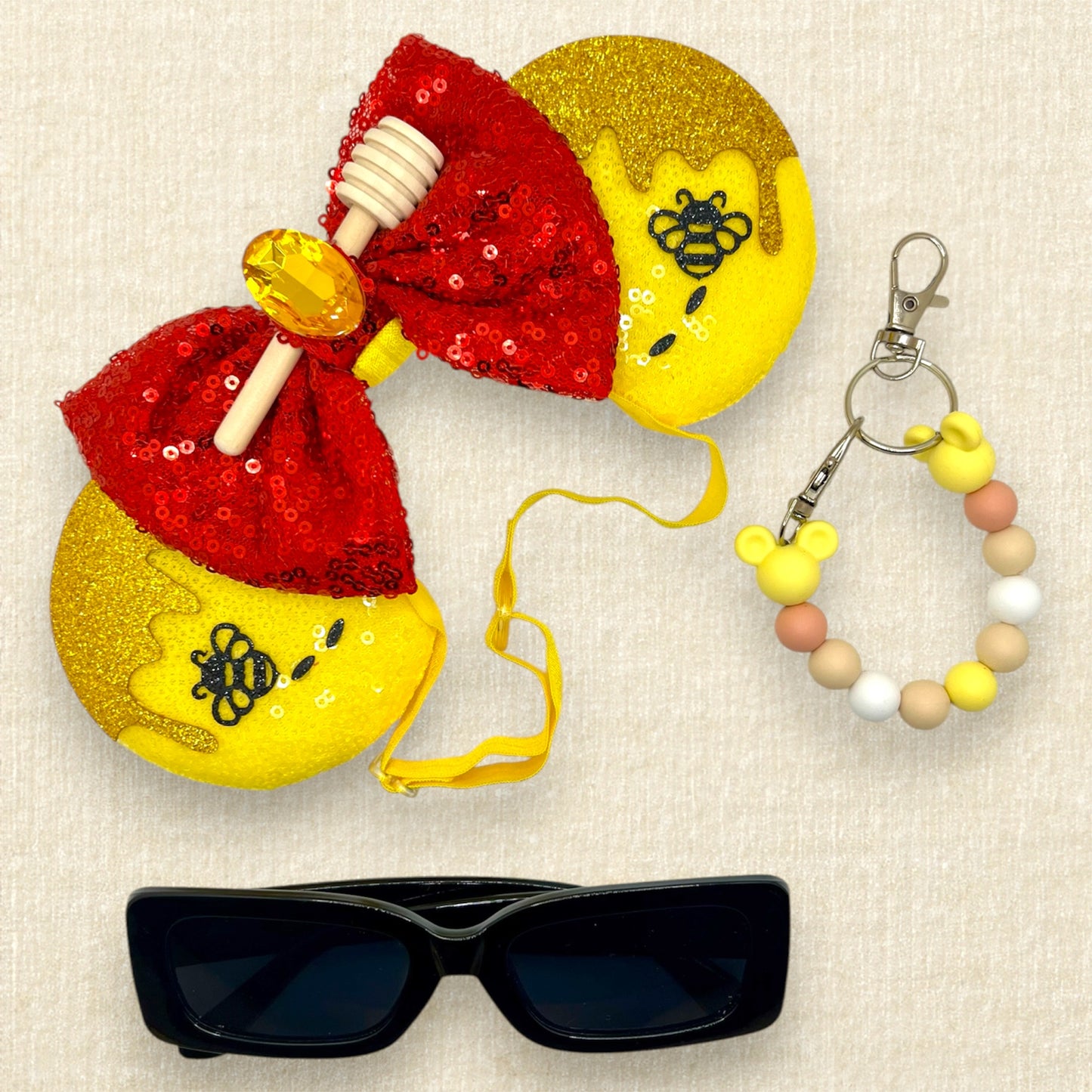 Yellow sequin mouse ears with glitter honey and bee accents, red sequin bow, wooden honey dipper, and yellow rhinestone on an elastic headband; black sunglasses; yellow beaded mouse ears holder keychain
