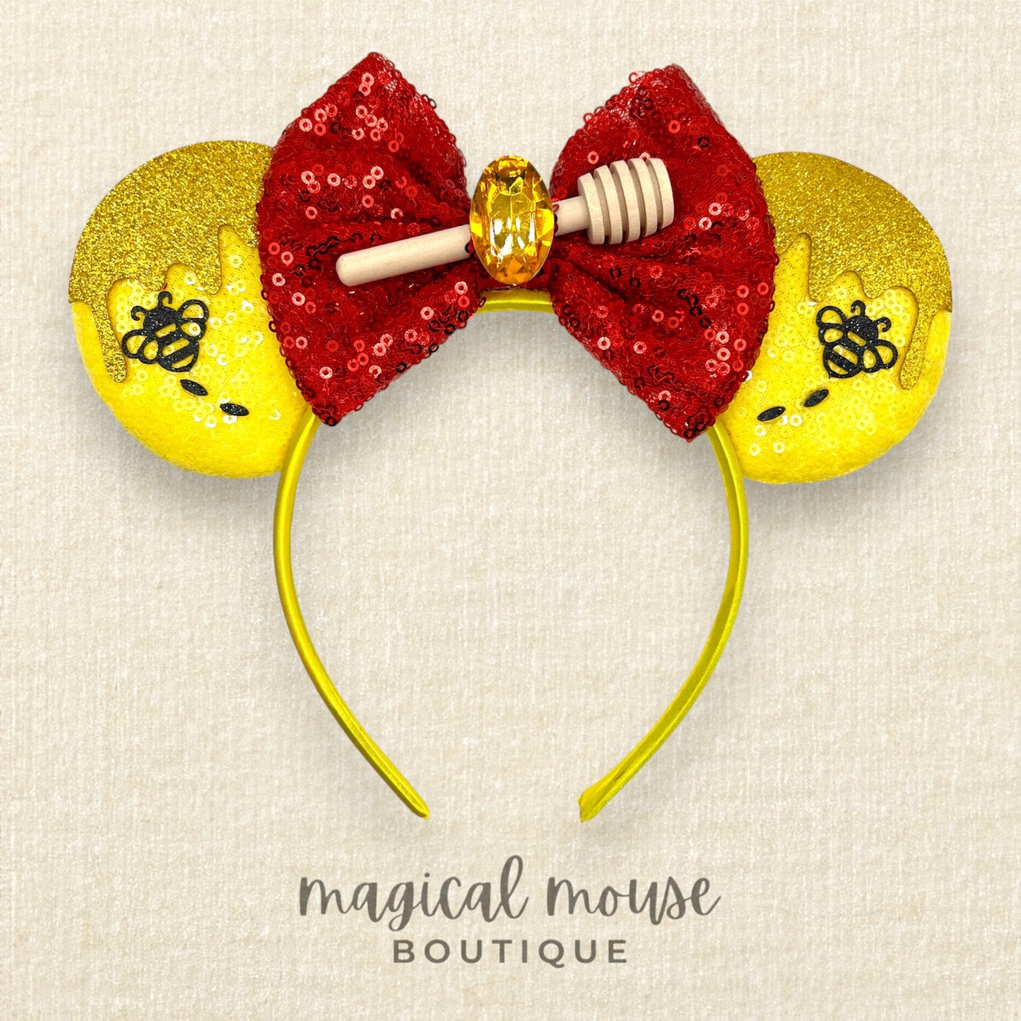 Mouse ears with a red sequin bow and yellow glittery ears with a honey and bee design on a beige background, branded 'magical mouse boutique'.