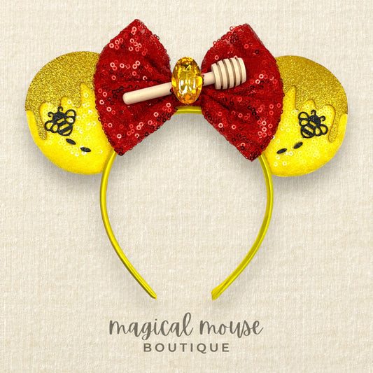 Mouse ears with a red sequin bow and yellow glittery ears with a honey and bee design on a beige background, branded 'magical mouse boutique'.