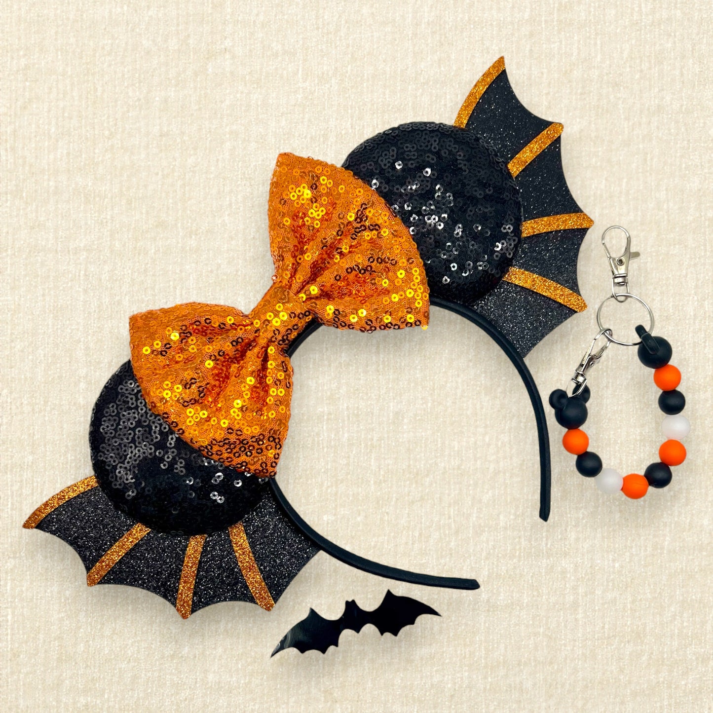 Halloween-themed headband with orange bow and black bat wings on a beige background; black decorative bat; orange and black beaded mouse ears holder keychain