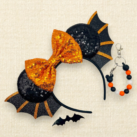 Halloween-themed headband with orange bow and black bat wings on a beige background; black decorative bat; orange and black beaded mouse ears holder keychain
