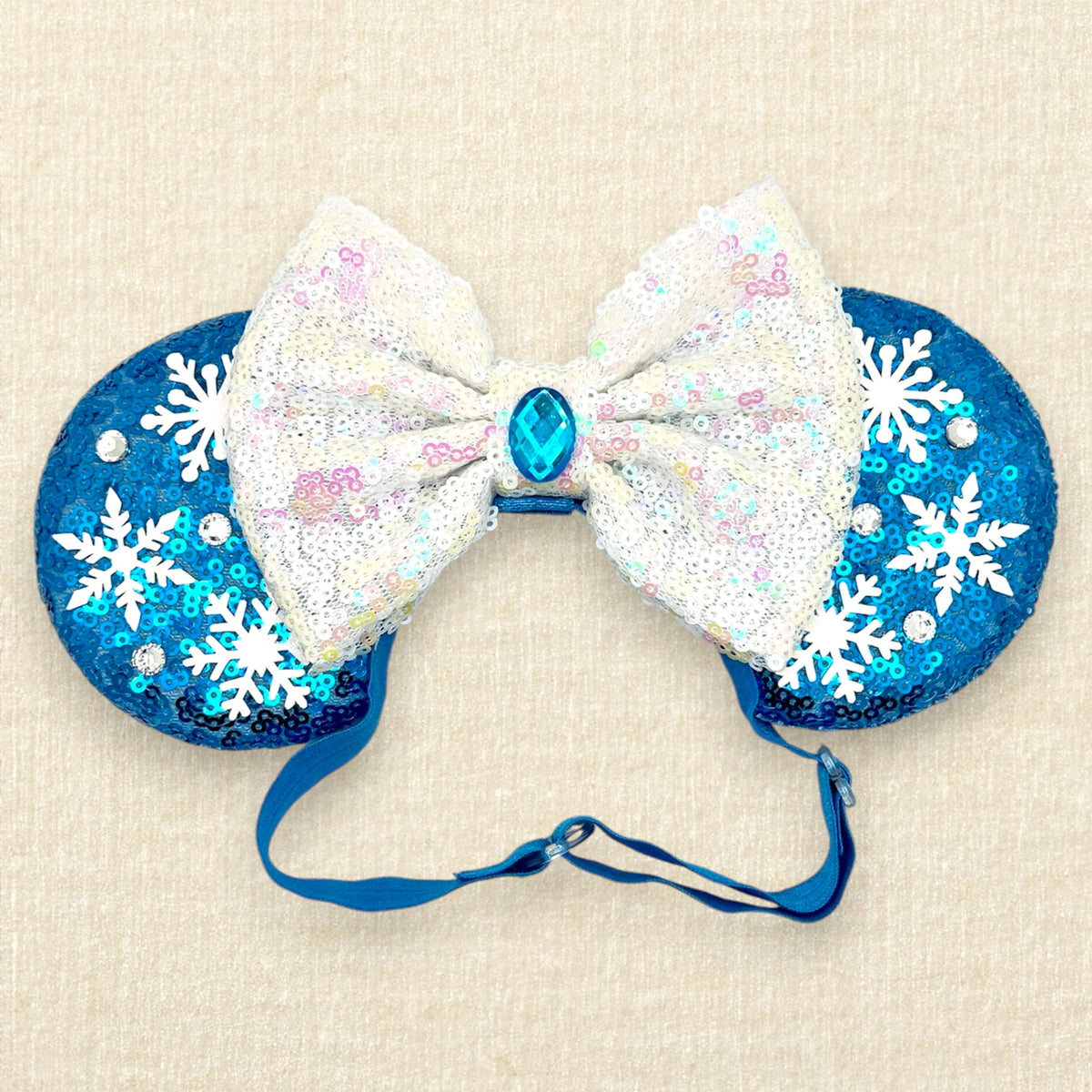 A pair of winter-themed blue sequined mouse ears with white snowflake embellishments and a large white sequined bow in the center with an elastic headband, set against a light beige textured background.