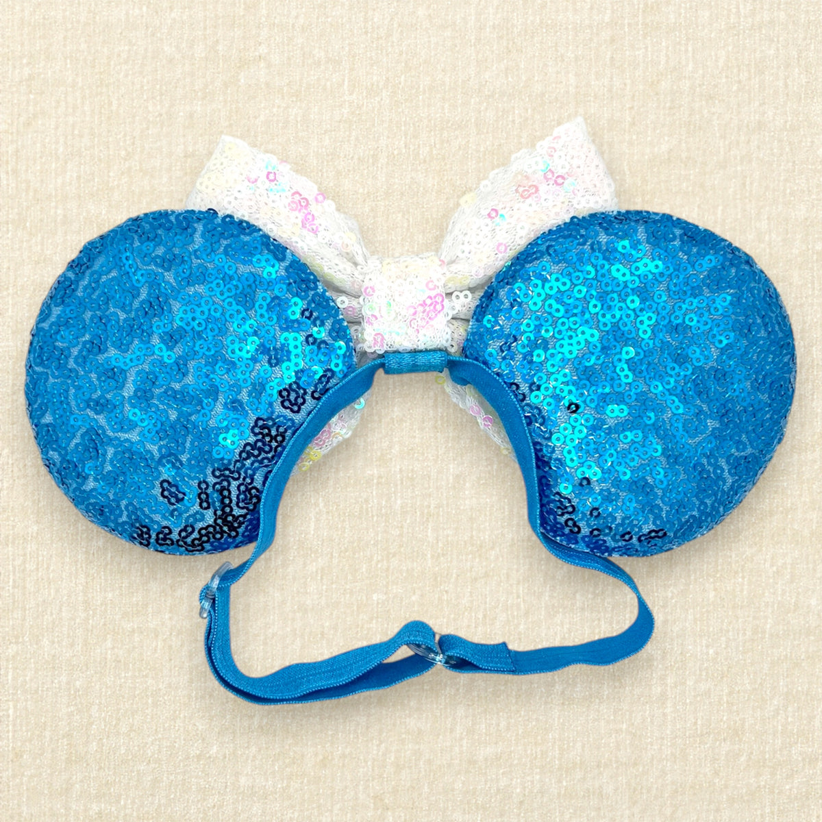 A pair of blue sequined mouse ears with white snowflake embellishments and a large white sequined bow in the center with an elastic headband, set against a light beige textured background (back view)