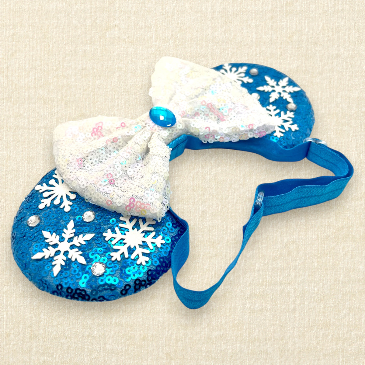 A pair of blue sequined mouse ears with white snowflake embellishments and a large white sequined bow in the center with an elastic headband, set against a light beige textured background.