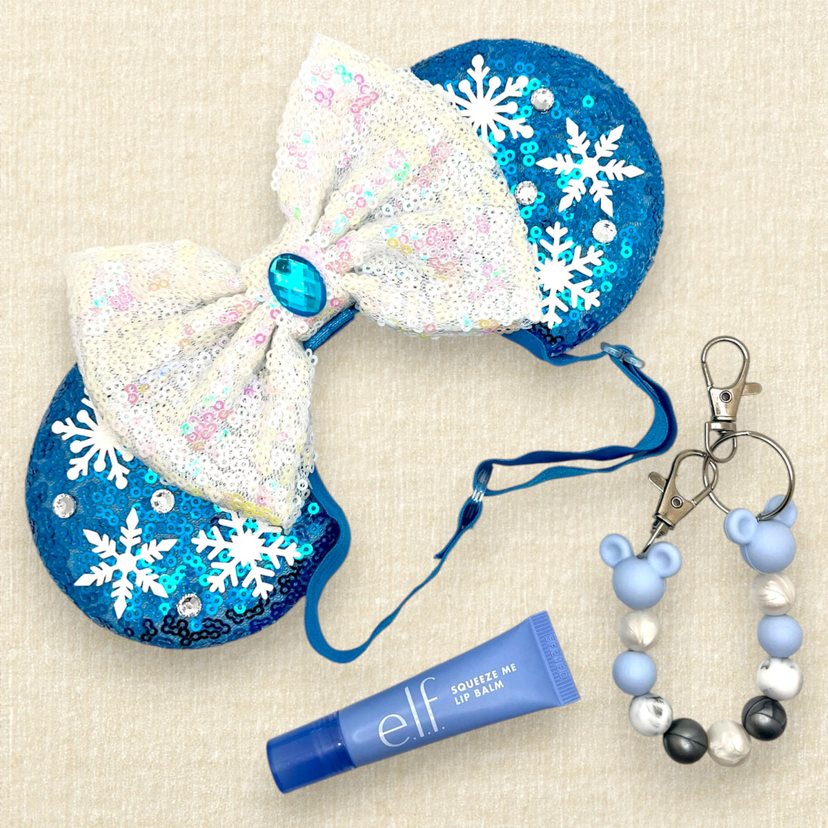 A pair of winter-themed blue sequined mouse ears with white snowflake embellishments and a large white sequined bow in the center with an elastic headband; a tube of blue lip gloss; and a blue, pearl white, and silver beaded mouse ears holder keychain clip